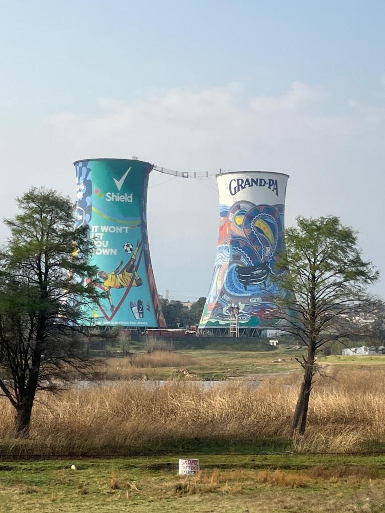 A view of two large, colorful cooling towers featuring murals, one with the text 'Shield' and the other labeled 'Grand-Pa', set against a cloudy sky.