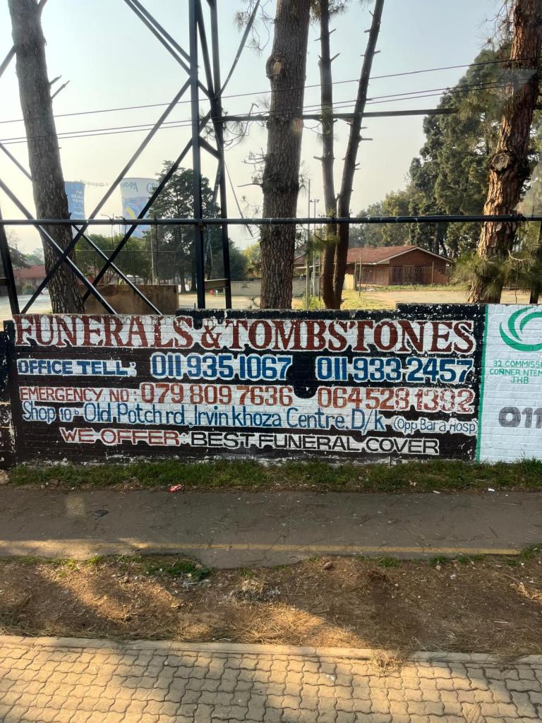 A roadside advertisement for a funeral and tombstone service, displaying contact numbers and an emergency number. The sign emphasizes offering the best funeral cover, located near Irvin Khoza Centre.