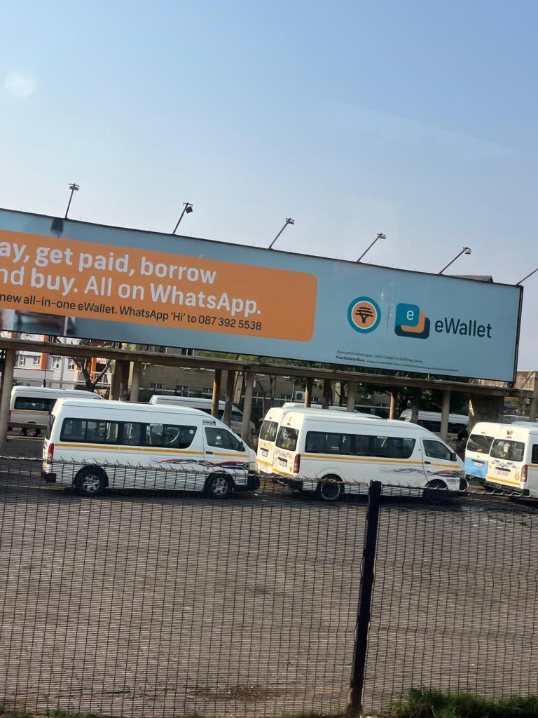 A large billboard advertising eWallet services, featuring messaging about payments and WhatsApp usage, above a parking area with several white vans.