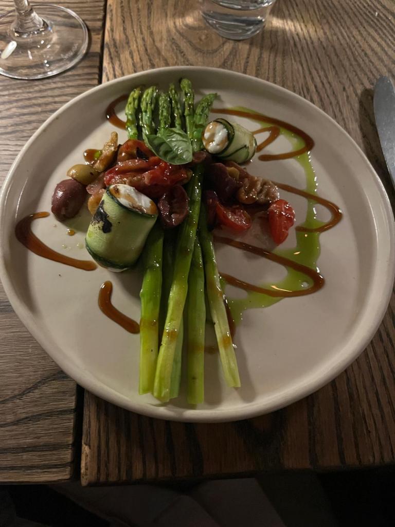 A plate featuring grilled asparagus topped with rolled zucchini filled with cheese, cherry tomatoes, olives, and walnuts, drizzled with balsamic glaze and olive oil.