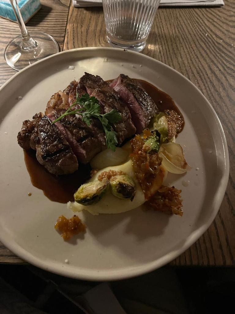 A close-up of a plate featuring a grilled steak, sliced to show the pink interior, garnished with a sprig of parsley. Accompanied by roasted Brussels sprouts and a sweet sauce.