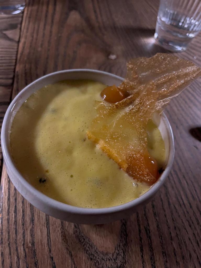 A small white bowl containing a creamy yellow dessert topped with a crisp caramelized piece and small orange spheres, resting on a wooden table.