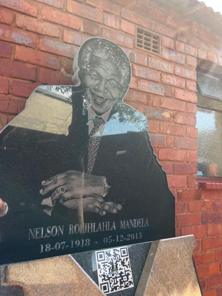 A stone memorial of Nelson Mandela featuring his smiling portrait and engraved details, set against a red brick wall.