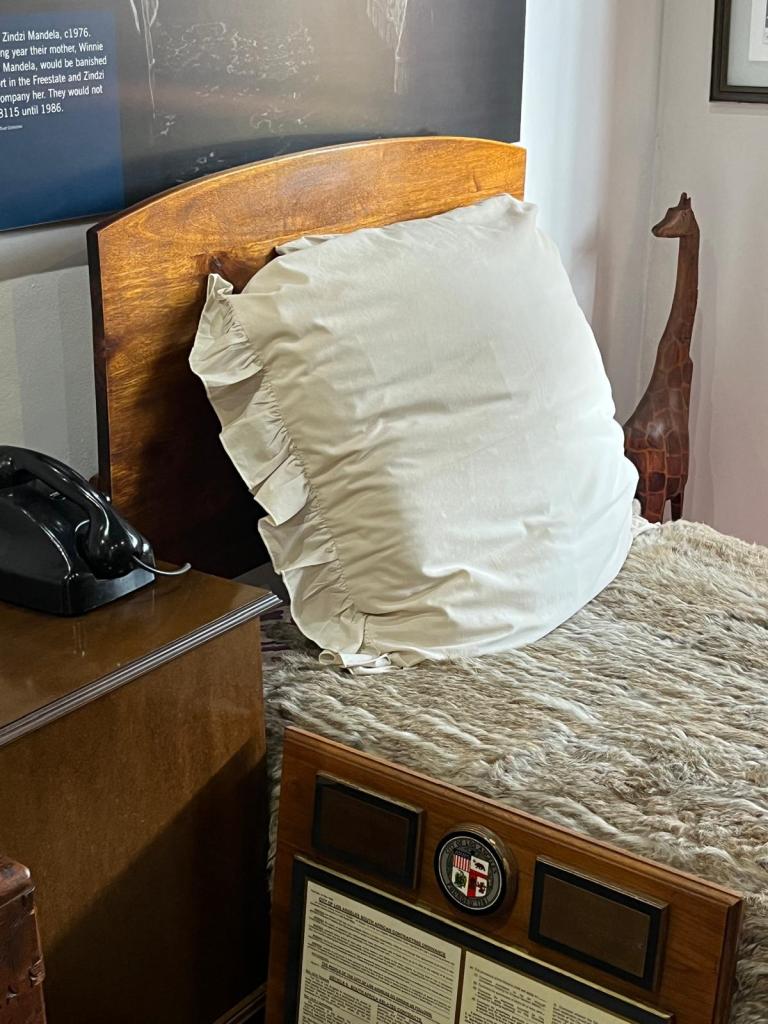 A cozy bed featuring a large white decorative pillow, a textured grey blanket, and a wooden headboard. Next to the bed is a vintage telephone and a display with a plaque.