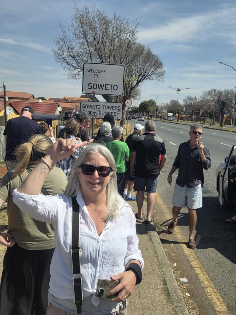 A group of people standing near a sign welcoming visitors to Soweto, with one woman in the foreground smiling and waving with her hand raised.