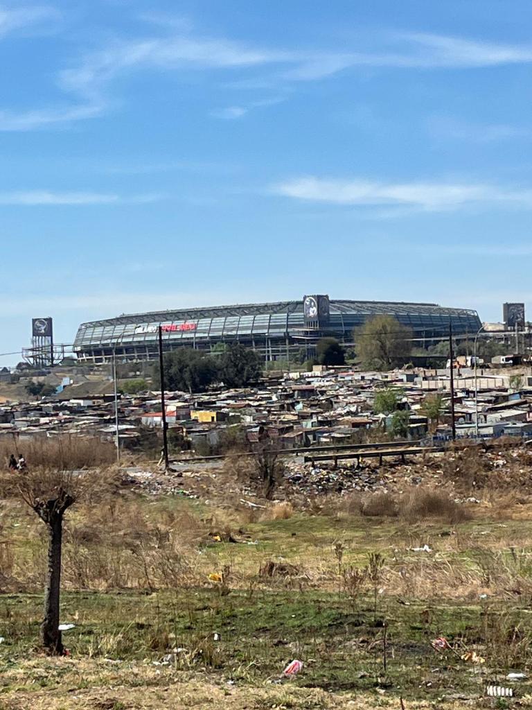 A view of a large stadium in the background, surrounded by a residential area with makeshift housing and scattered debris in the foreground under a clear blue sky.