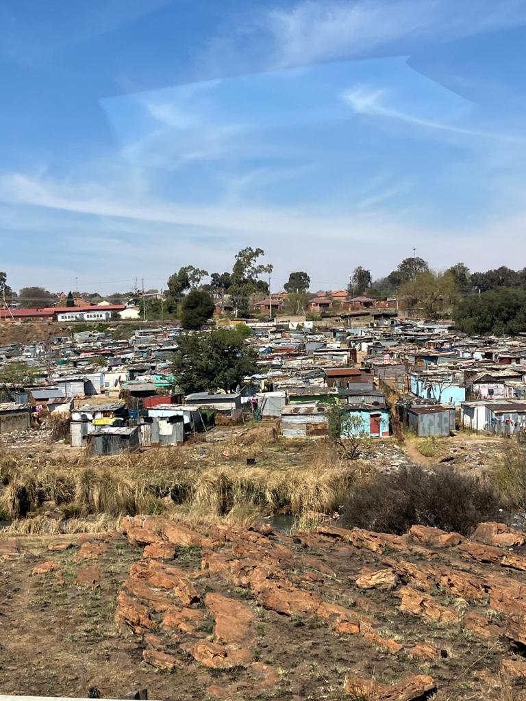 A view of a densely populated informal settlement with makeshift homes, set against a backdrop of clear skies and greenery.