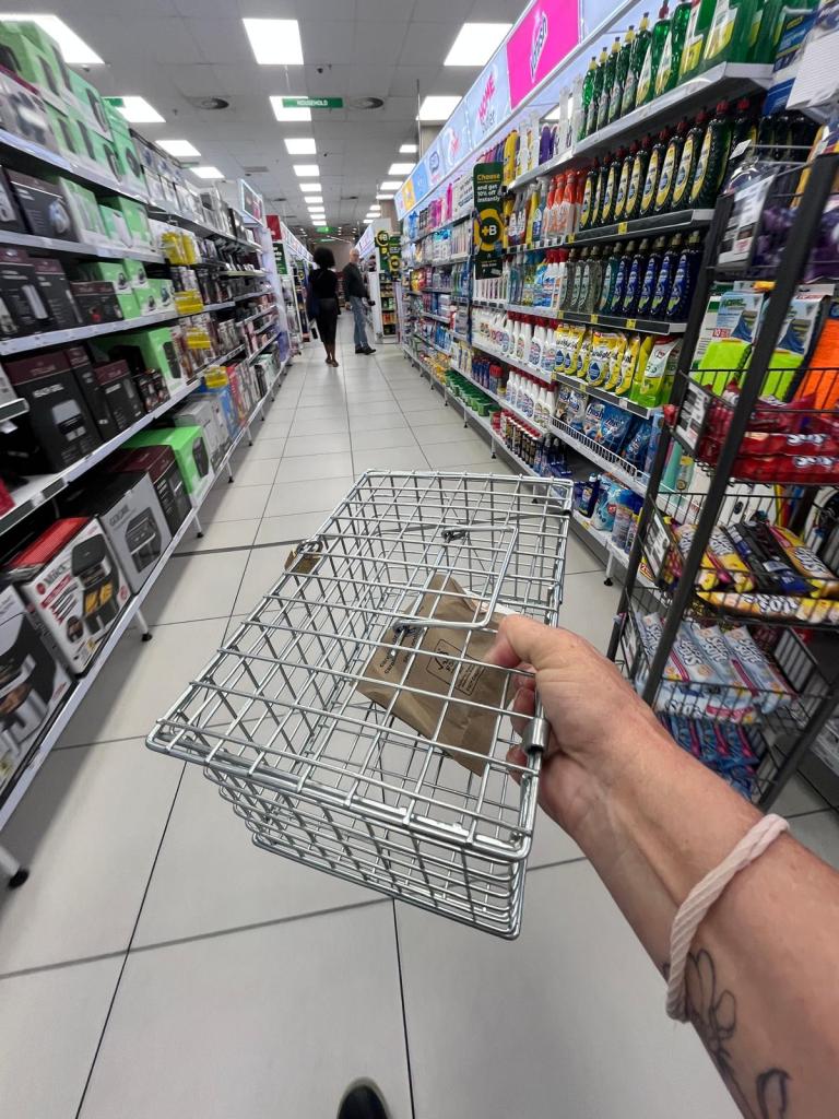 A person holding a metal shopping basket while walking down the aisle of a retail store, surrounded by shelves stocked with various products.
