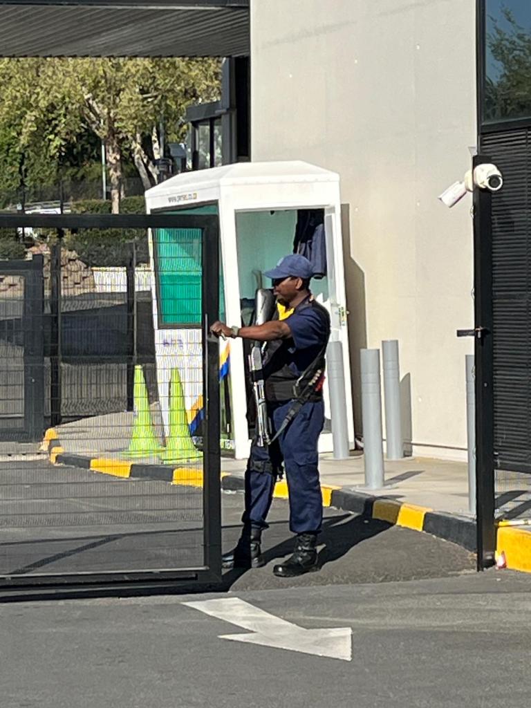 A security guard with a rifle stands at a gated entrance, partially opening the gate. The background features a secure booth and green traffic cones.